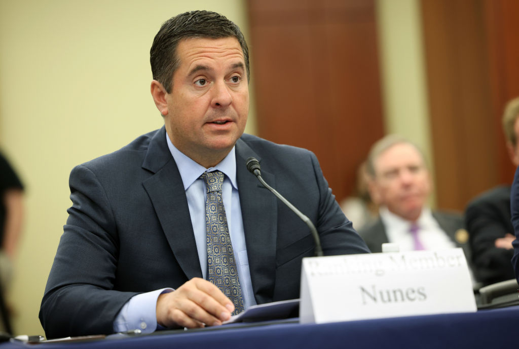 A man wearing a business suit sits at a table with a microphone in front of him and a sign that says "Nunes."