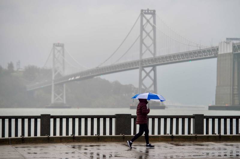 The person holds an umbrella and walks by themselves. The Bay Bridge and Yerba Buena Island are visible in the background but the heavy rain makes them hard to locate.