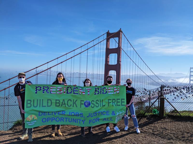 Five people wearing masks hold up a green sign that reads "President Biden Build Back Fossil Free" with a bridge in the background.