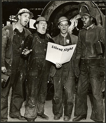 Four men wearing coveralls and hardhats sing the song Silent Night.