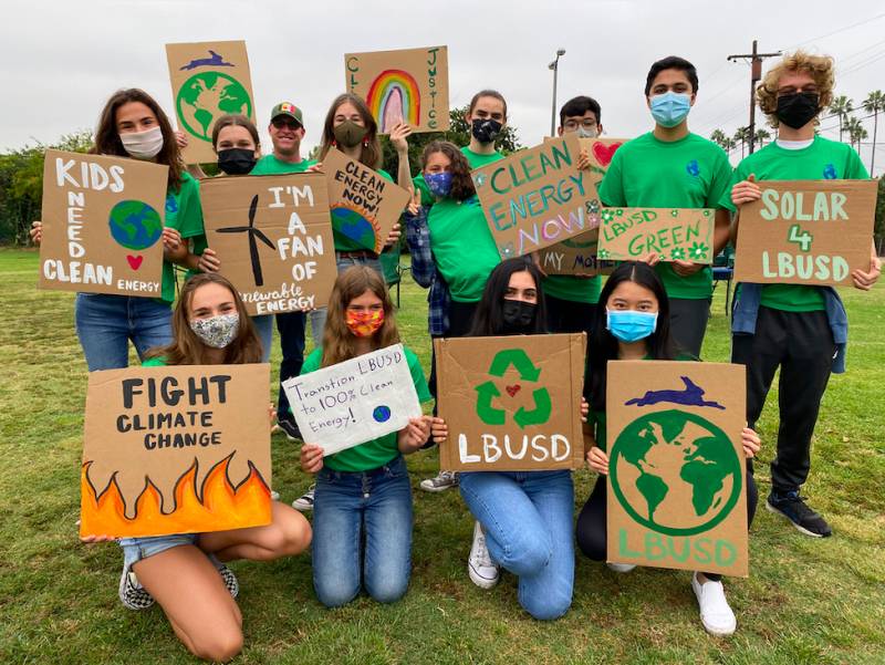 Thirteen people are kneeling and standing wearing green shirts, face masks and holding signs that read "Fight climate change" and "Solar LBUSD."