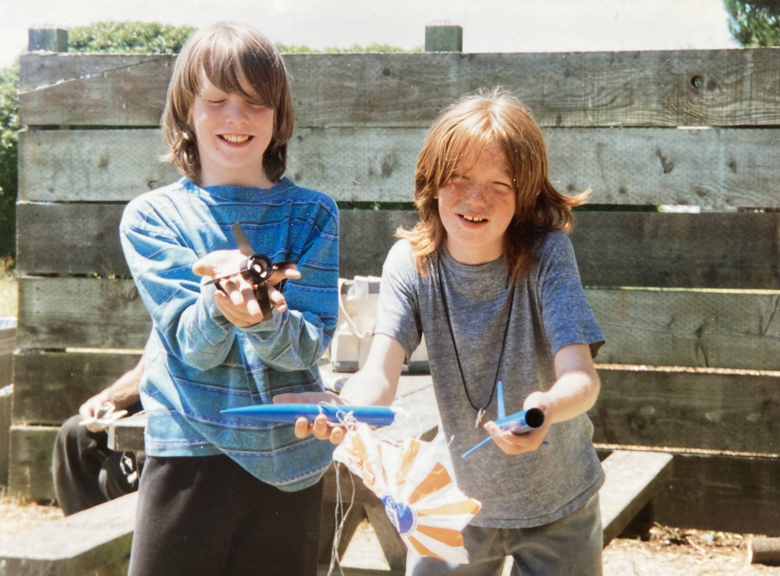 Two boys grin as they show off toy airplanes in an outdoor setting.