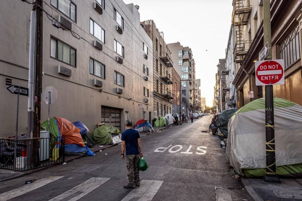 A person stands in the middle of the street and looks down the sidewalk where there are dozens of tents lined up