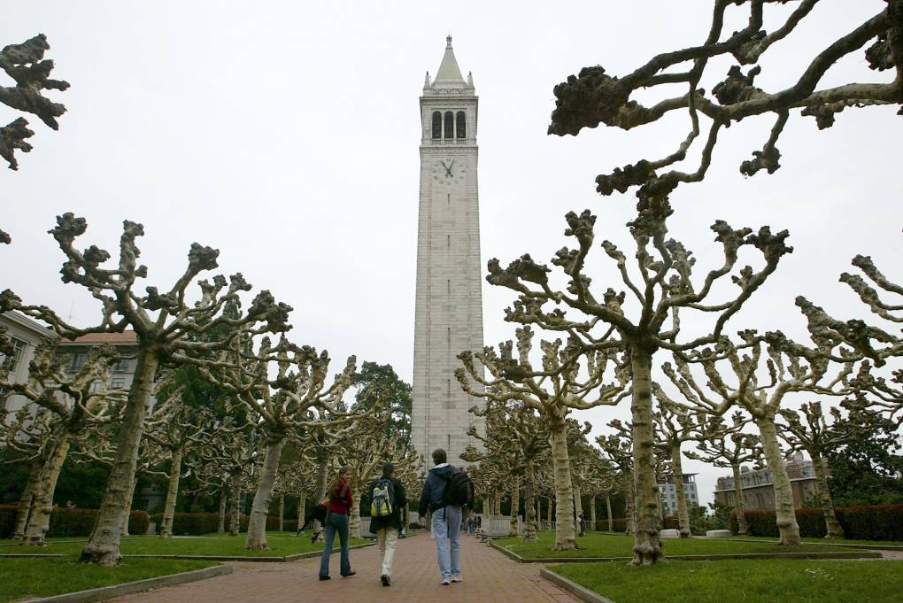 Three people are walking towards a tall building with a clocktower surrounded by trees with no leaves.