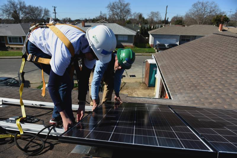 two workers on a roof in hard hats lean over a solar panel on a sunny day