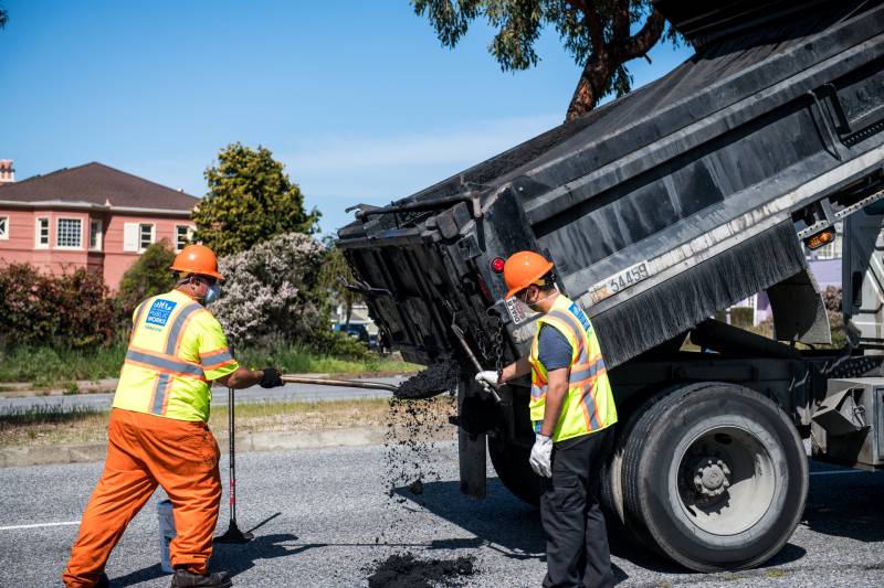 Two people wear neon vests, face masks and a helmet as they move asphalt from a truck to the street.