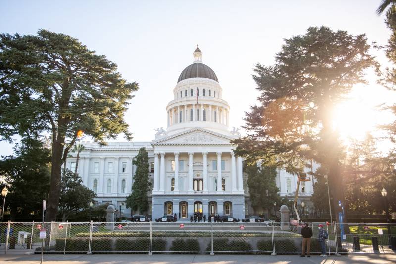 The California capitol building in the middle of the frame, with sunlight peaking through a tree on the right side with a fence in the foreground.