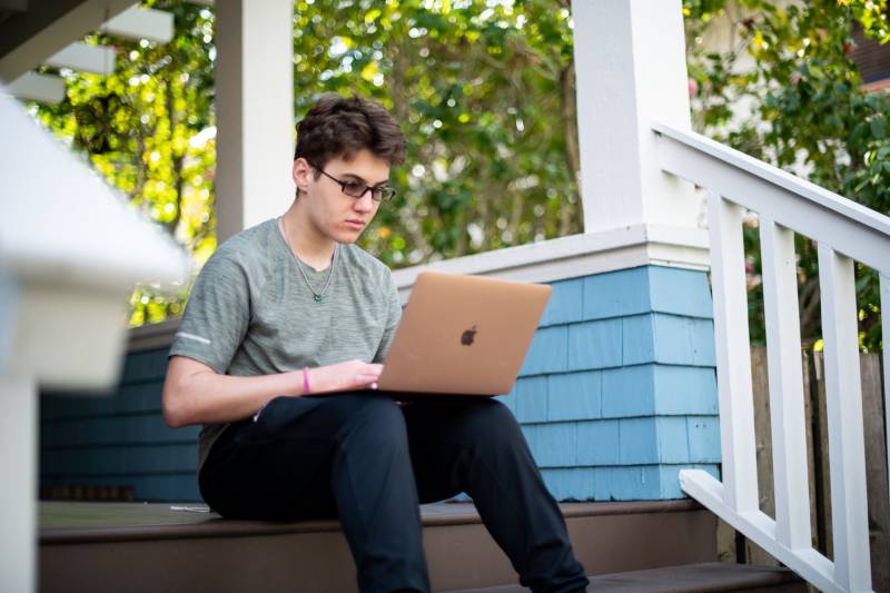 A young man wearing glasses and a grey t-shirt sits on the steps of a house with a laptop in his lap.