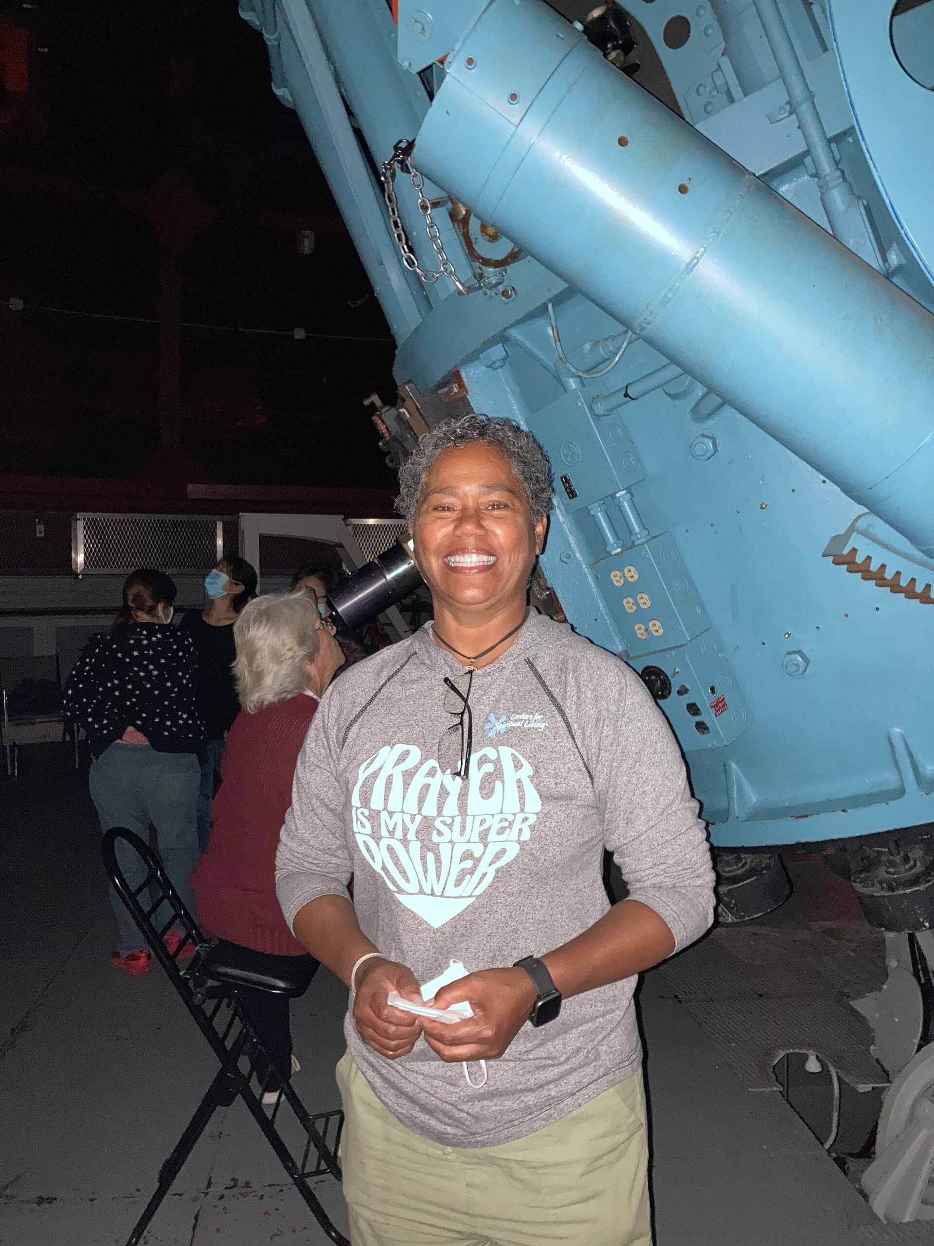 A person stands in front of a very large telescope and holds their face mask in their hands.