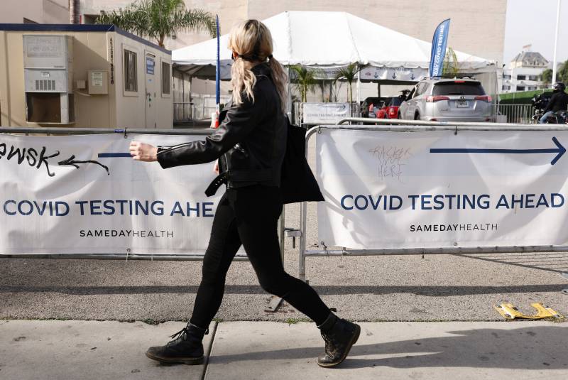 A women in black jeans and a black jacket walks past a white sign that says "COVID testing."