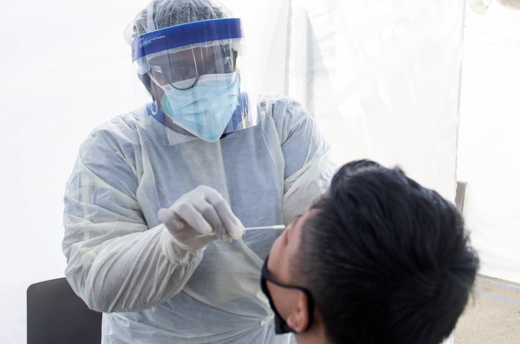 A man wearing a white plastic suit and gloves, with a surgical mask under a plastic face shield affixed with a blue headband, points a swap into the throat of a second person, with short black hair and a black face mask, pulled to their chin.
