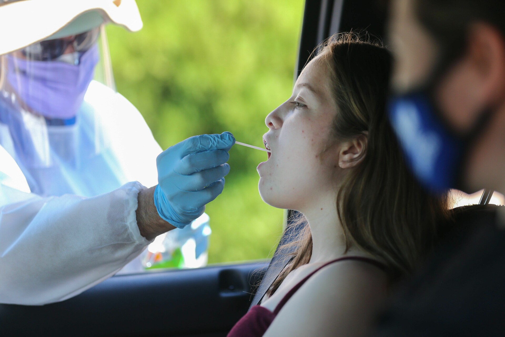 A healthcare worker wearing PPE reaches through an open car window to administer a throat swab to a girl sitting in the car.