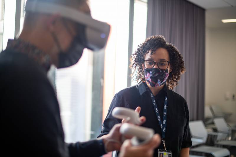 Two people stand in a room: one wears a virtual reality headset and holds two controllers while the second figure stands by and watches over.
