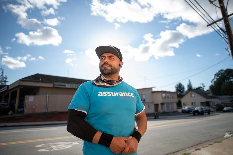 A man stands outside holding his hands wearing a hat and blue shirt that says "Assurance."