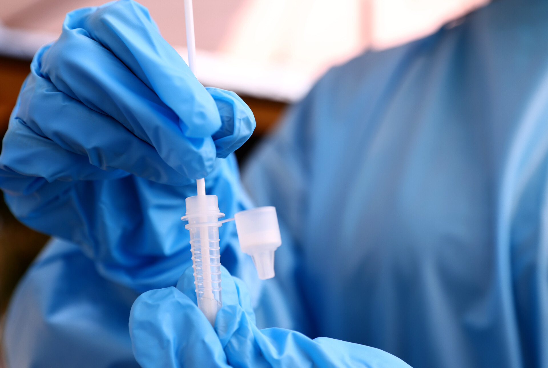 A gloved blue hands held by a nurse in a blue robe hold a small white COVID-19 testing vial and swab.