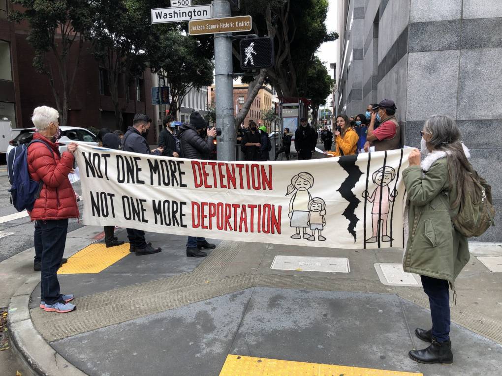 Two women hold a banner on a street corner that says 'Not One More Detention, Not One More Deportation.'