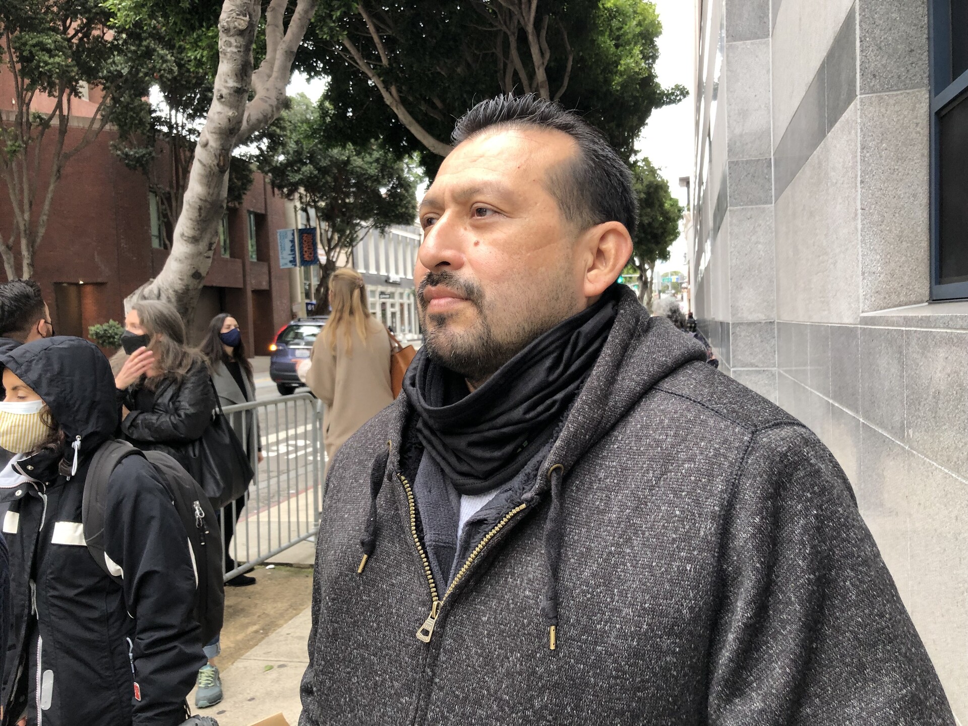 A man wearing a jacket and a scarf on a street in San Francisco, with a small group of protesters behind him.