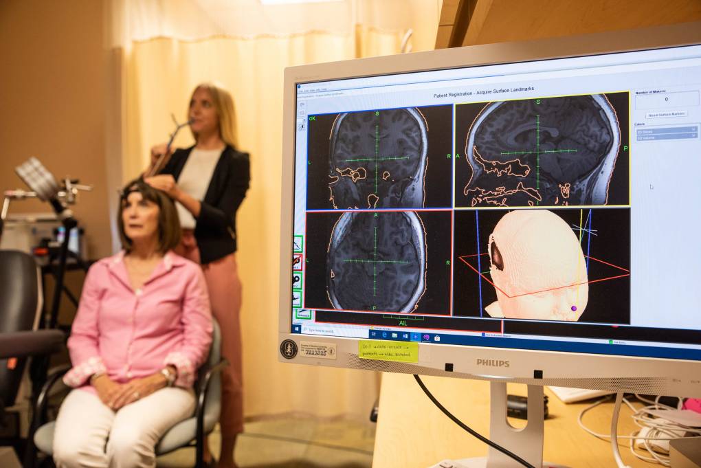 A computer monitor shows four panels of the human brain with two women in the background. One woman is wearing a pink shirt and sitting while a woman stands behind her holding a thin metal object to the top of her head.