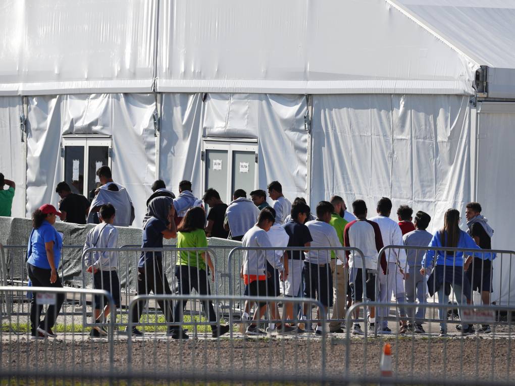 Children walk in a line past barricades toward a large white tent.