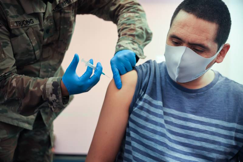 A man wearing a white mask and blue t-shirt has a needle pushed into his arm by a person wearing military fatigues and blue surgical gloves.