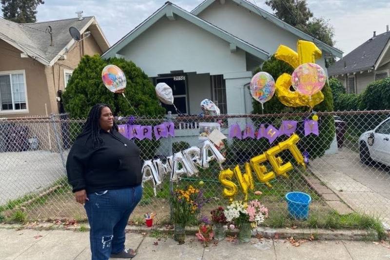 A woman wearing a black hoodie and jeans stands outside of a house near a fence with flowers, balloons and message that reads "Happy Birthday Shamara sweet."