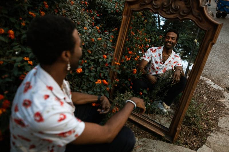 Smiling person in a white floral shirt smiles at themself in a mirror next to a bush filled with red flowers. 