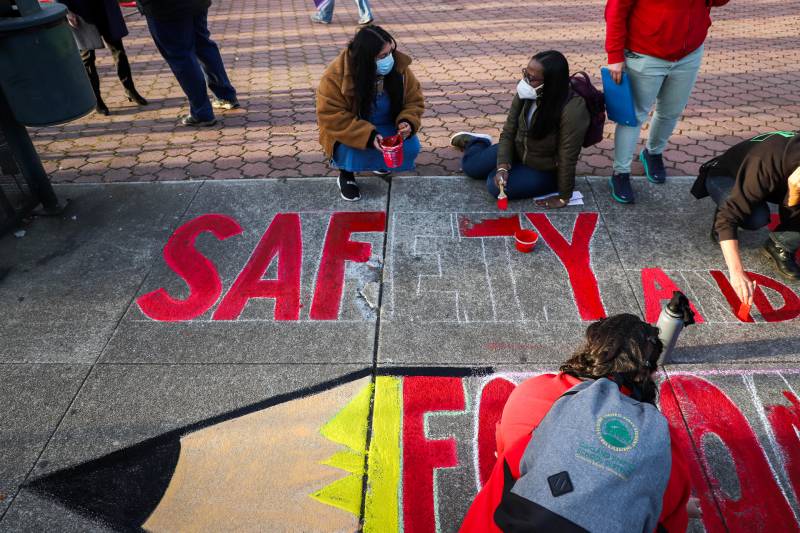 Three people wearing masks are seen writing words in chalk on the ground outside spelling "safety."