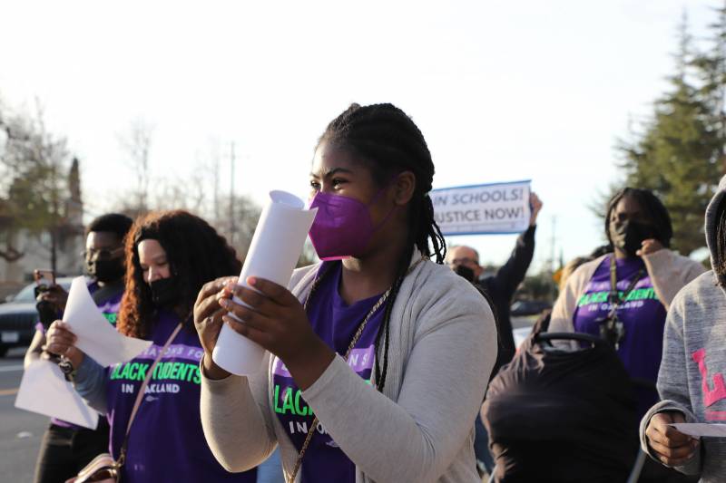 A woman with a purple mask and t-shirt holds a piece of paper rolled up in her hand with others in the background.