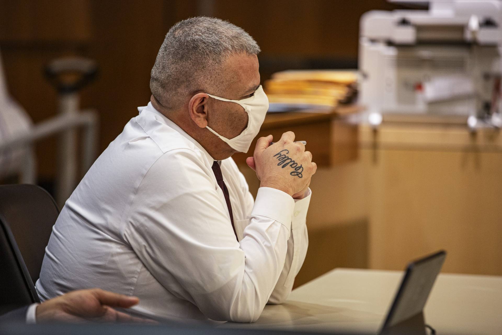 A man wearing a white button down shirt and mask is sitting with his elbows on a desk.