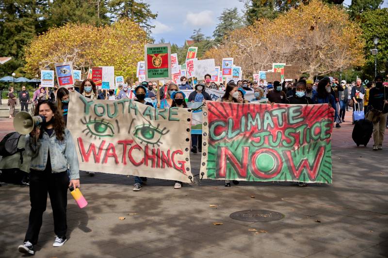 A march with several people wearing masks and holding signs that read "Youth are Watching" and "Climate Justice Now."