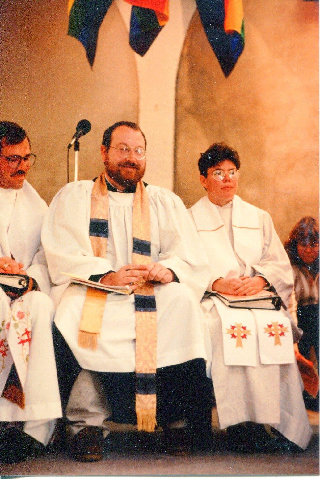 Three pastors wearing church garb sit near a microphone.