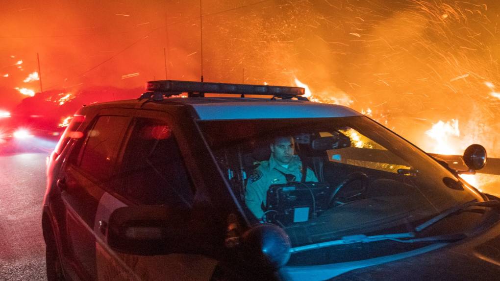A CHP officer seen cast in blue, sitting in the front of his SUV, while a wildfire blazes behind him.