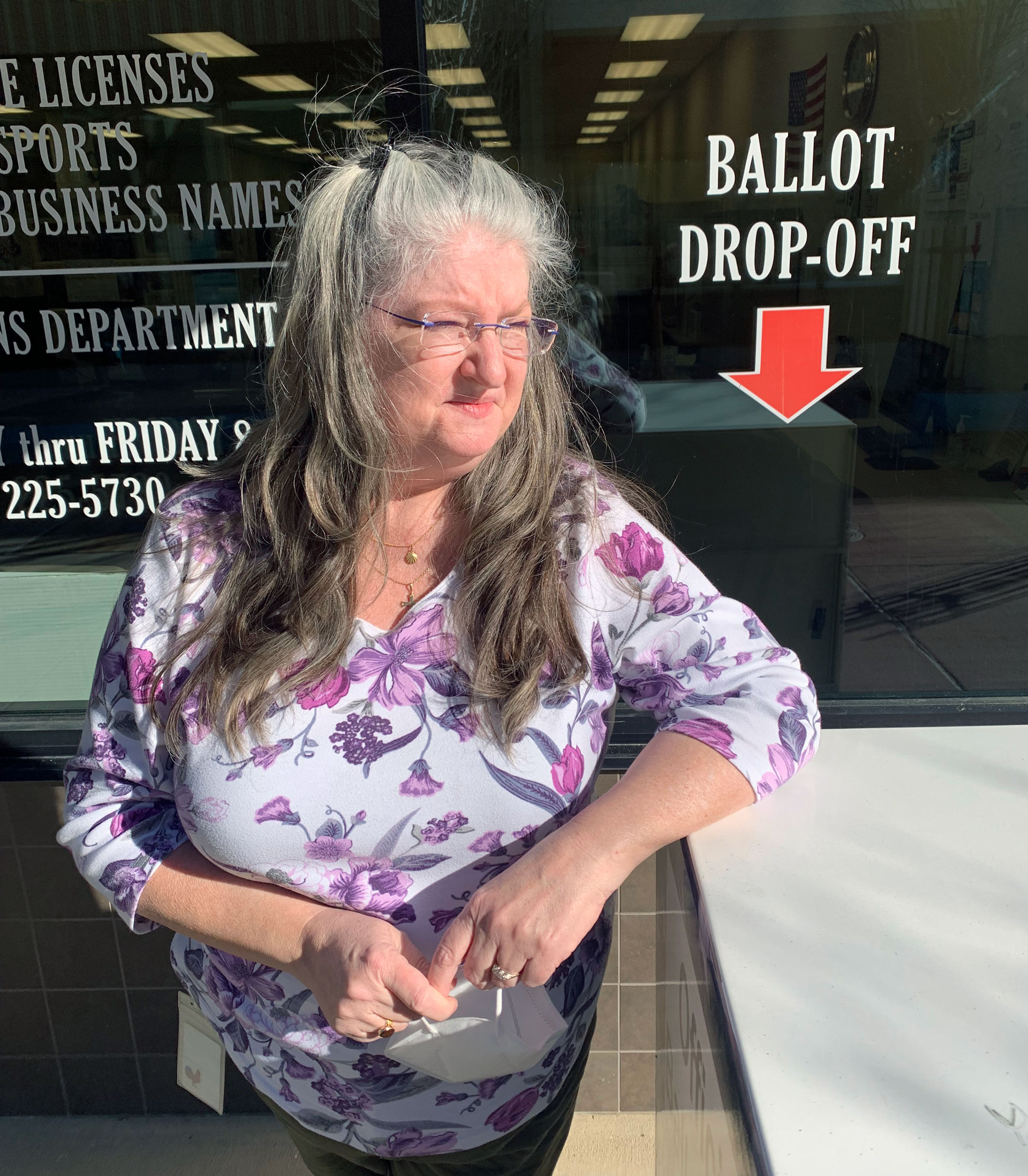 Woman stands outside, squinting, in front of window of county building, with 'ballot drop off' sign visible