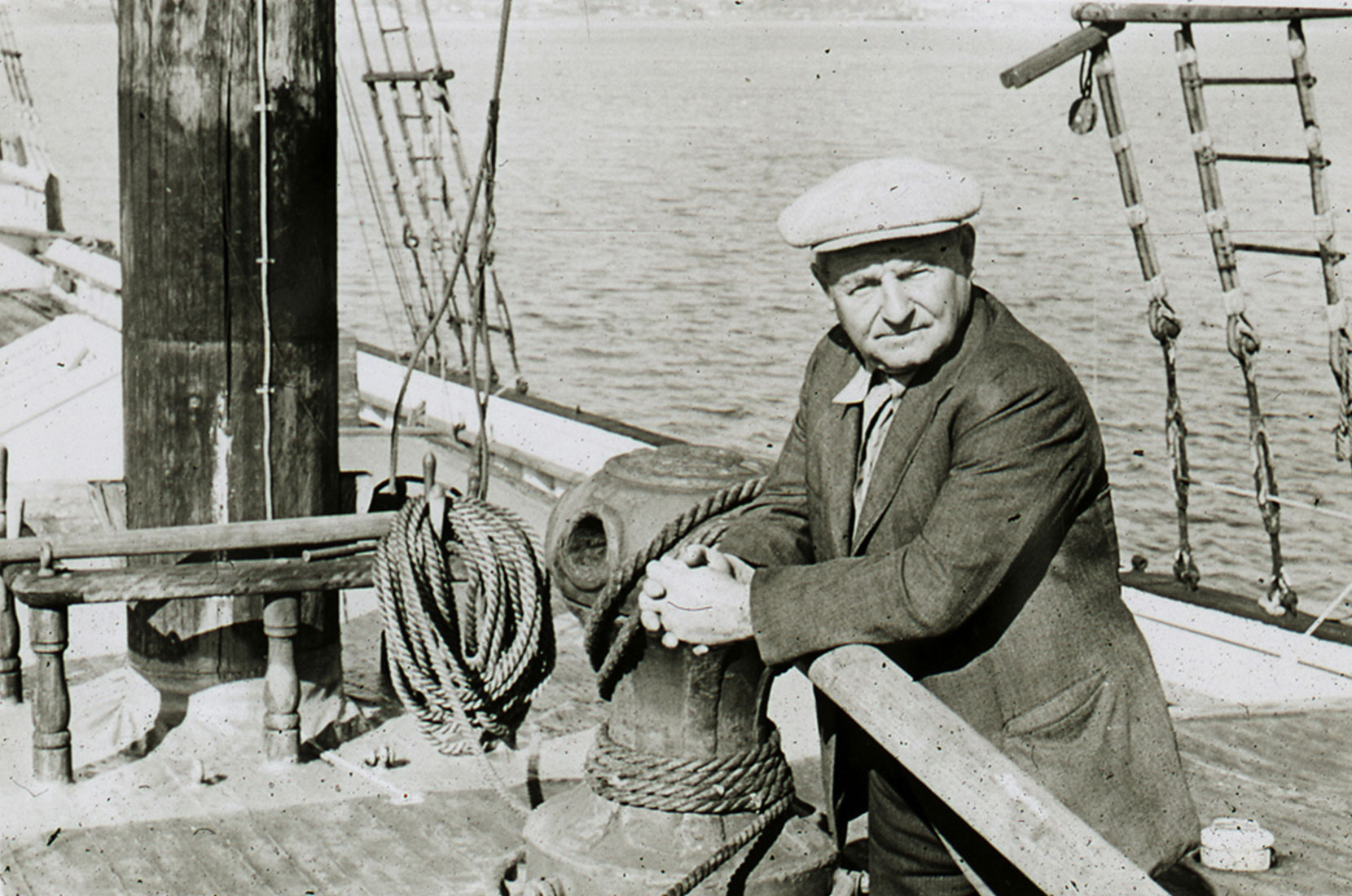 Black and white photo of a man in a worn sport coat and newsies cap leaning on the rail of a ship. A mask and rigging can be seen in the background.