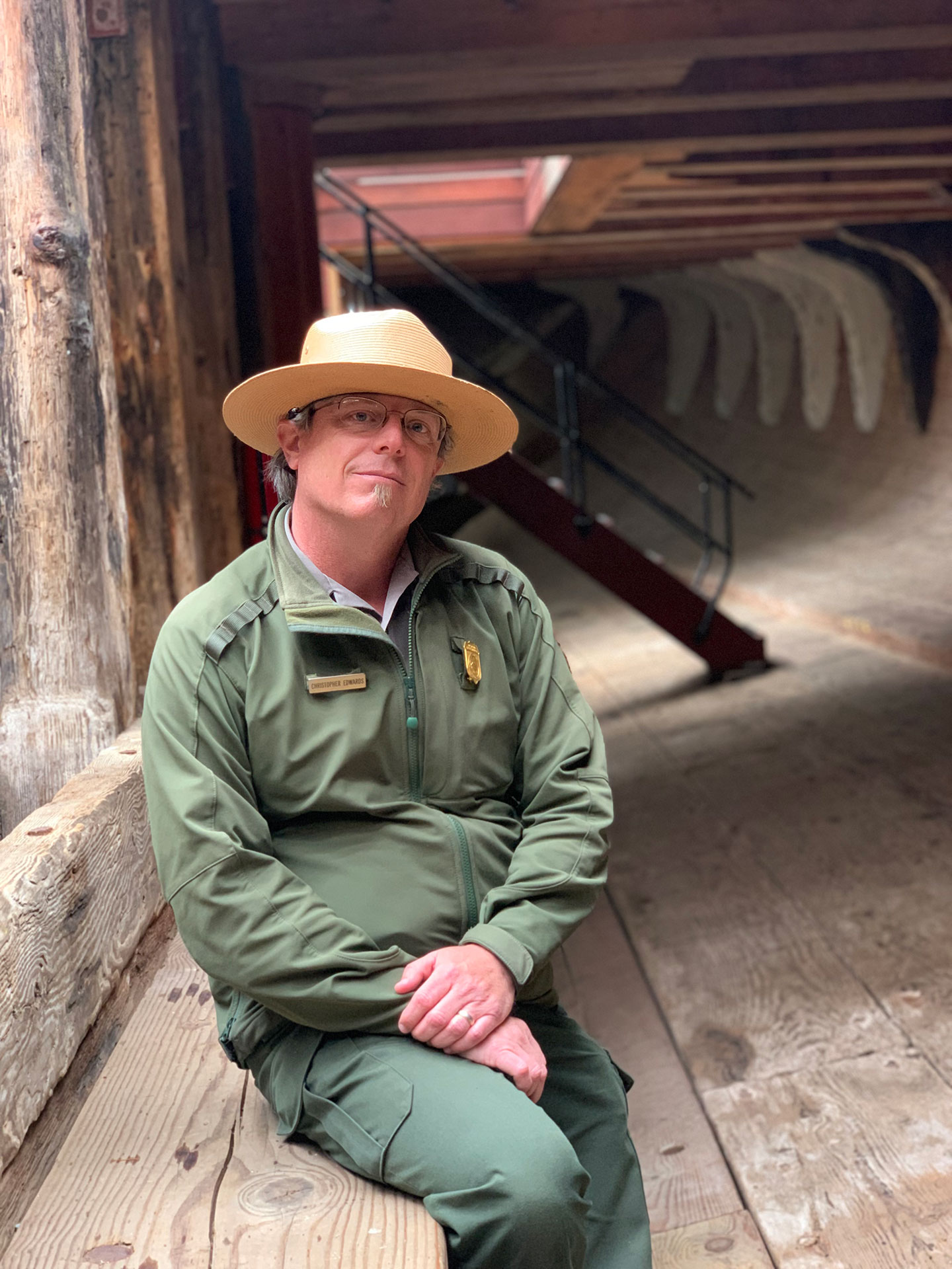 A man in a green park ranger uniform with a brimmed hat poses for a picture sitting in the hold of a large wooden ship.