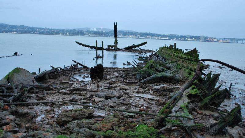 In the foreground is a muddy pile shaped vagule like the hull of a boat. Ropes lie on the ground and what looks like the remains of a bow can be seen. In the background, across the water, is a shoreline.