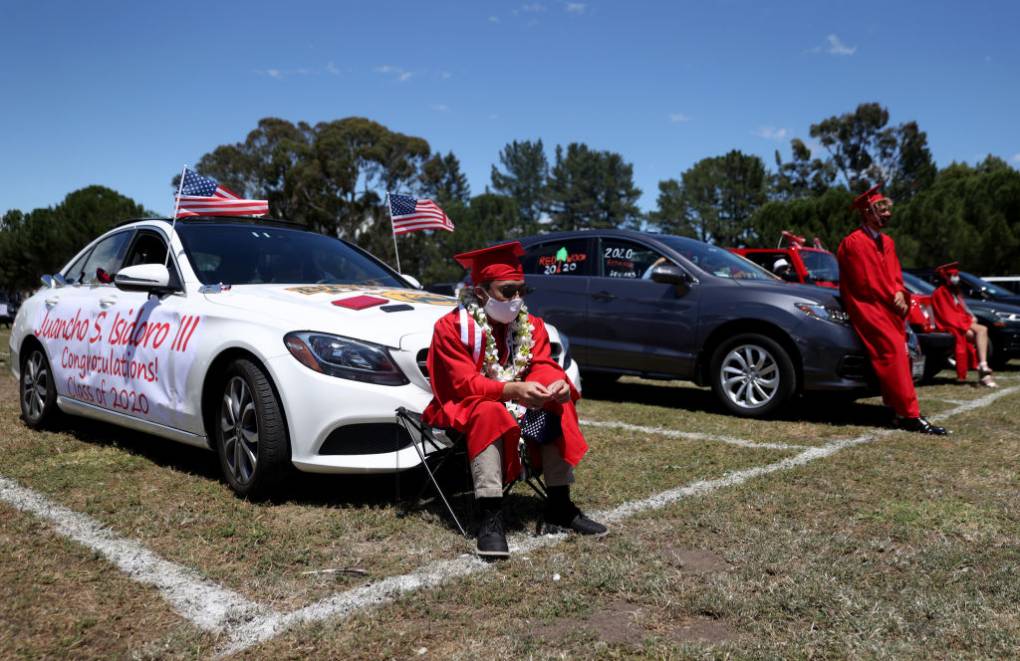 A young man dressed in a red cap and gown with black shoes, dress pants, sun glasses and a mask sits in front of a white car next to other people similarly dressed in front of cars in a field.