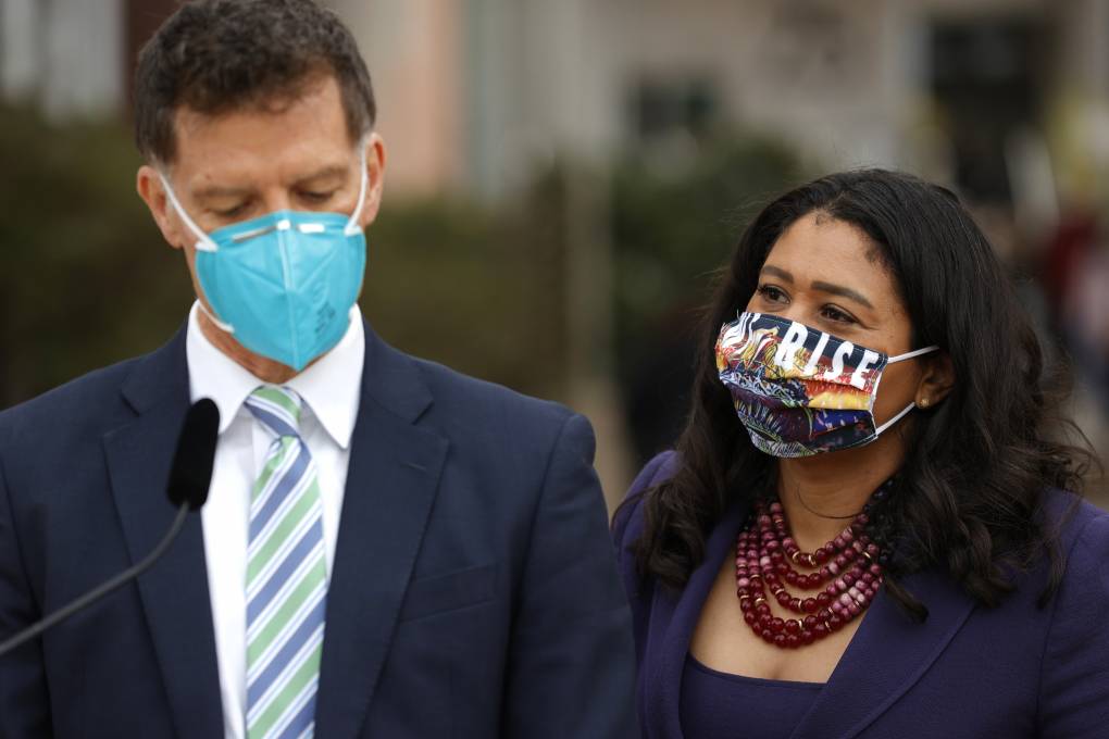 A man and woman stand next to each other. The man is wearing a business suit and light blue mask in front of a microphone and the woman is wearing a navy blue jacket with a red beaded necklace and multicolored mask.