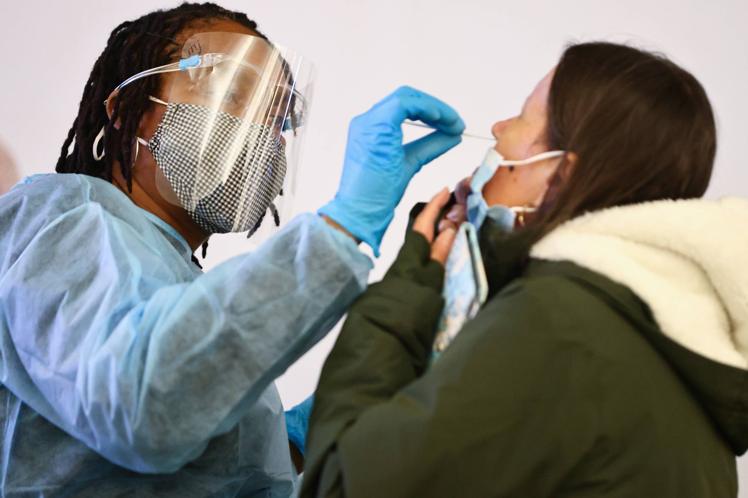 A person wearing scrubs and a face mask and a plastic protector sticks a nose swab into another person's nose.