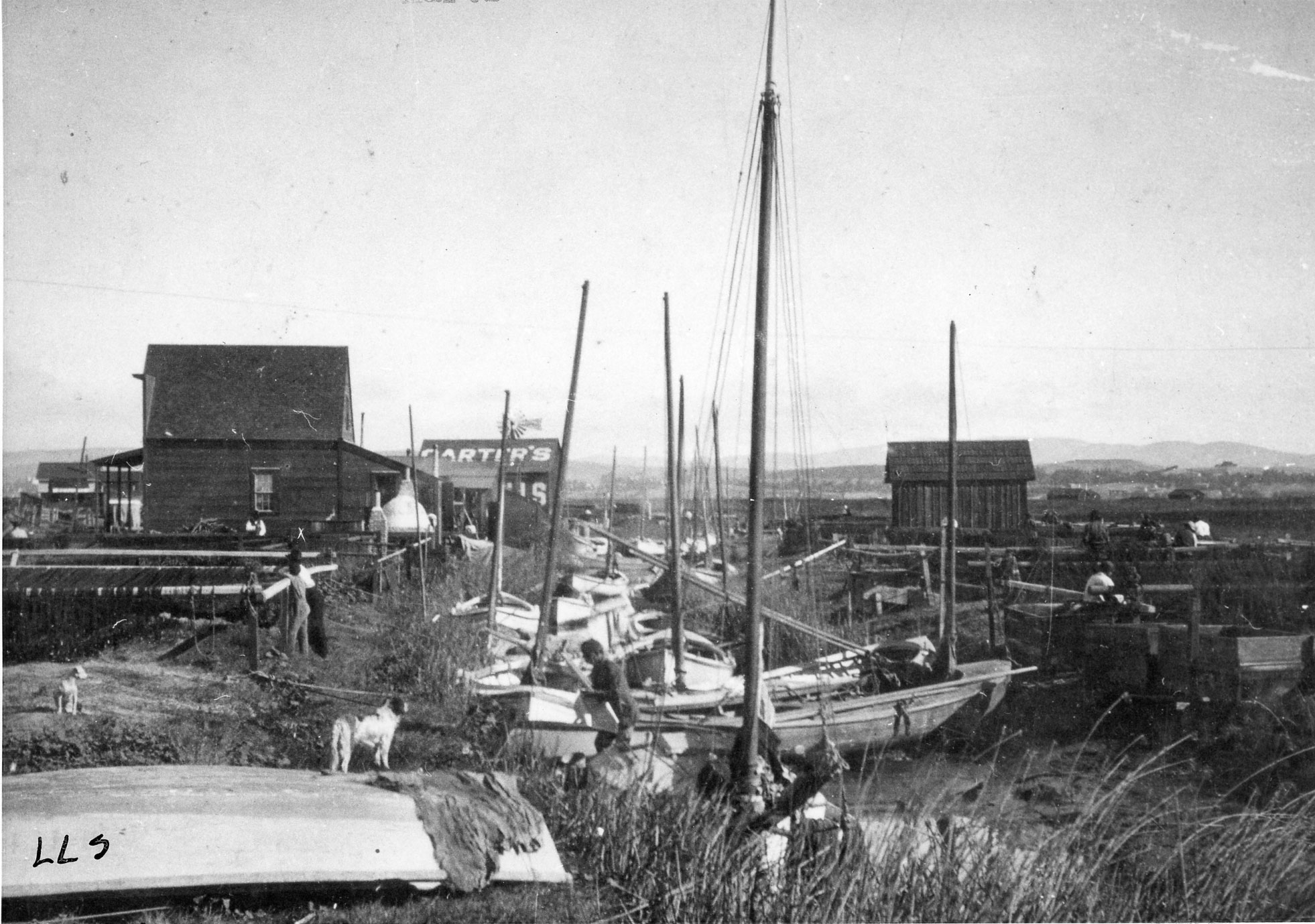 Black and white photo of fishing boats tied up in a creek with rustic buildings built in a marshland.