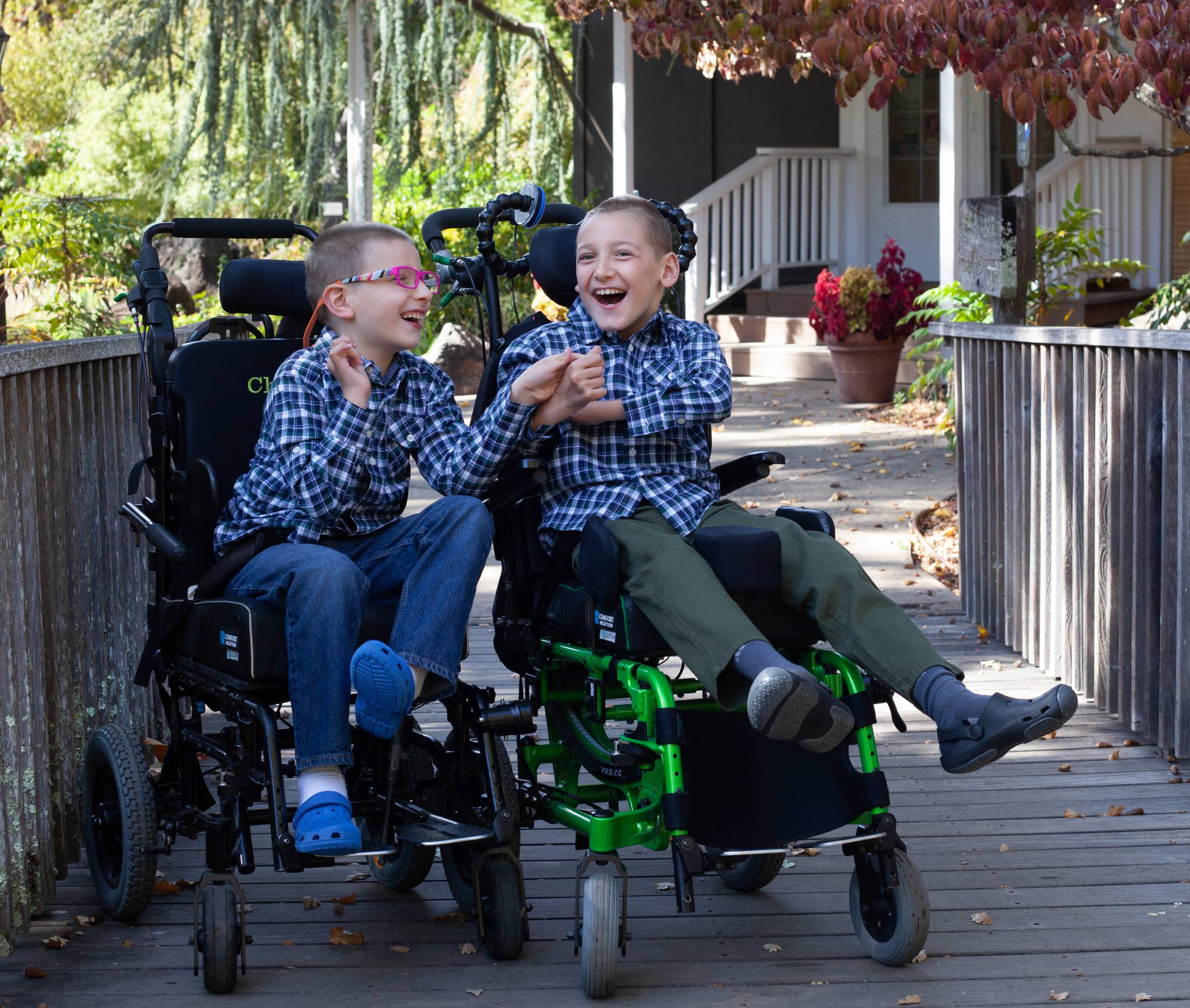 Two boys in wheelchairs, smiling and touching hands.