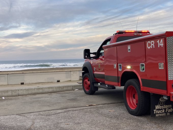 An emergency vehicle, red, sits on the right side of the frame facing Ocean Beach which features stormy waves.