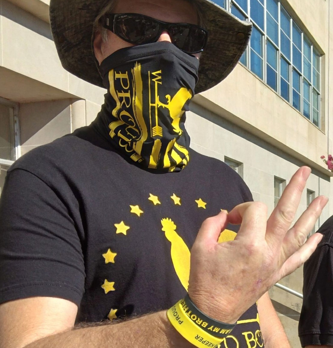 man wearing sunglasses and mask and yellow and black 'proud boys' t-shirt makes the 'ok' symbol