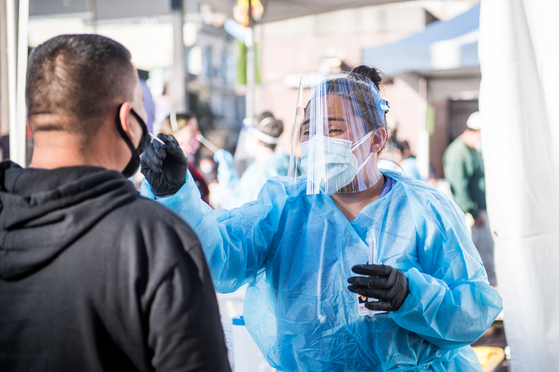 nurse in full PPE prepares to swab man wearing a mask facing away from camera, outside in sunlight