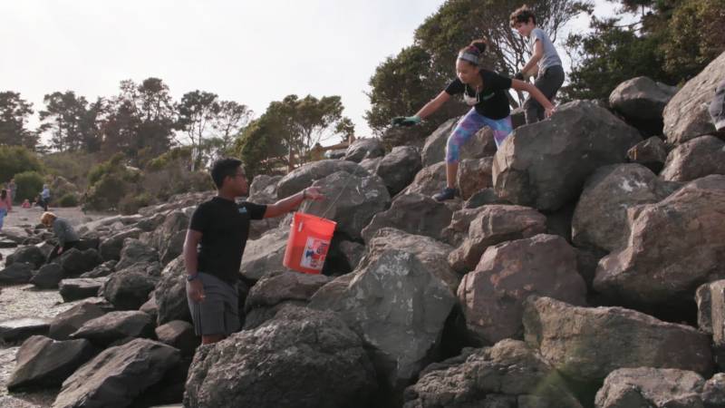 Three young people stand on a rock pile. One hands an orange bucket to another.