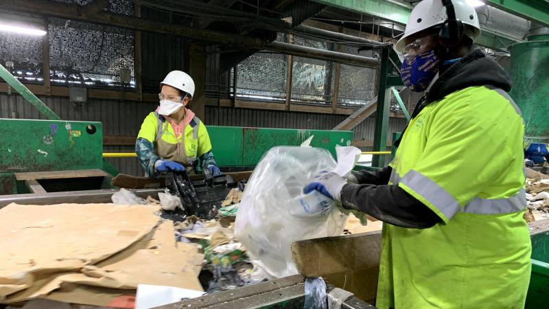 Workers in green safety jackets and helmets sort trash along a conveyor belt. 