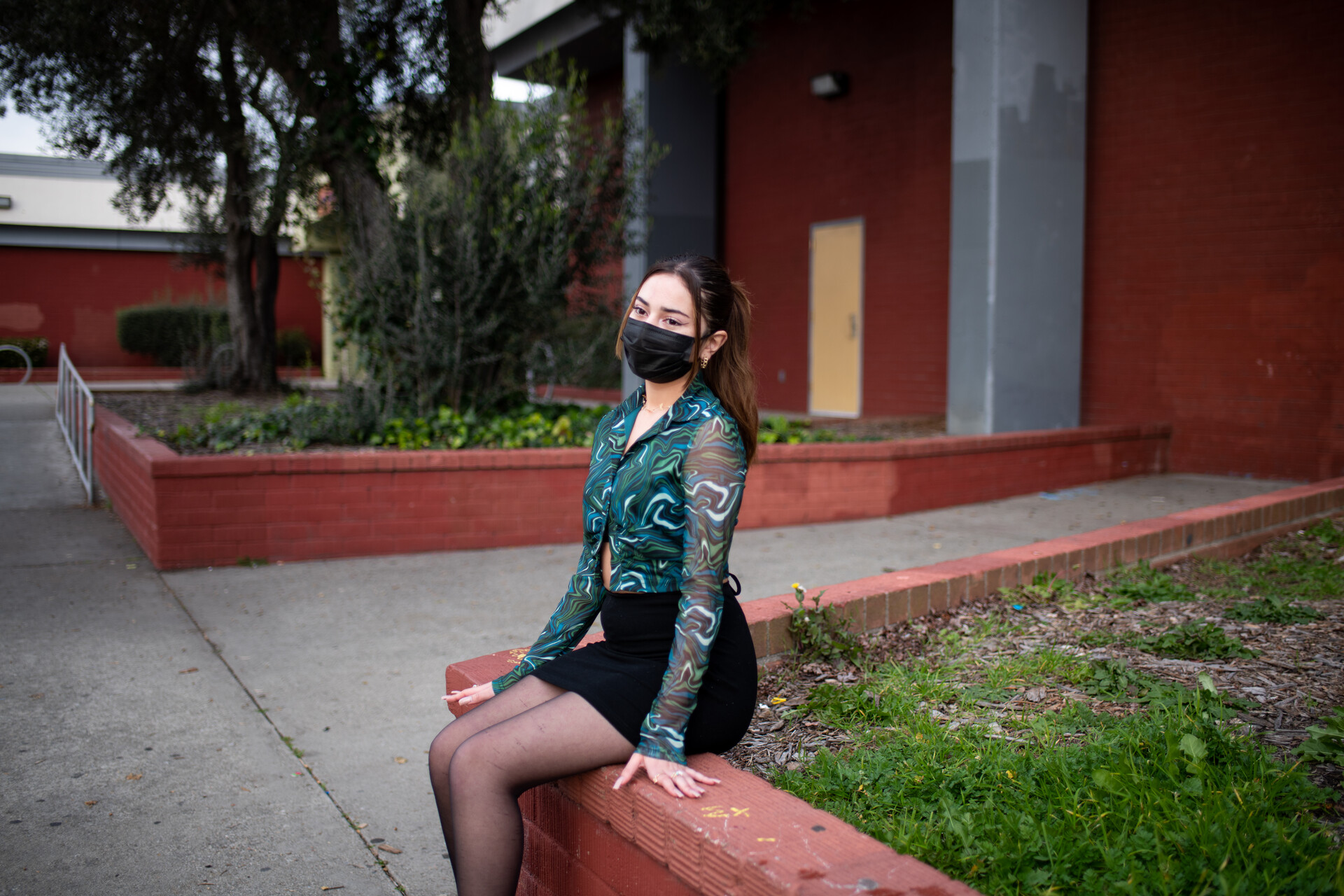 young girl wearing mask sits with pensive look outside an entrance to her high school