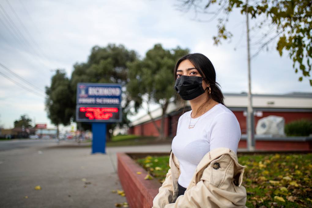 Teenage girl wearing mask looks toward camera, with sign reading Richmond High School in background