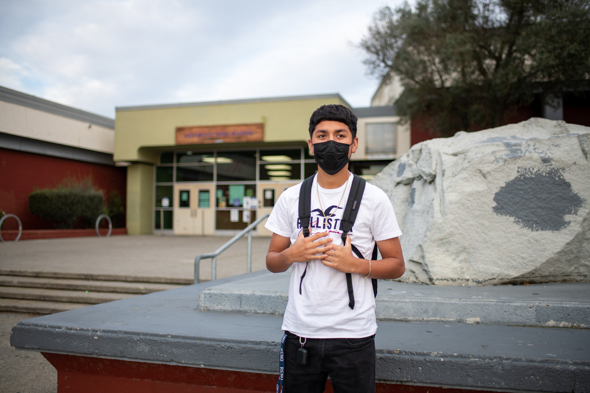 male high school senior wearing mask and white t shirt stands with a backpack in front of high school entrance