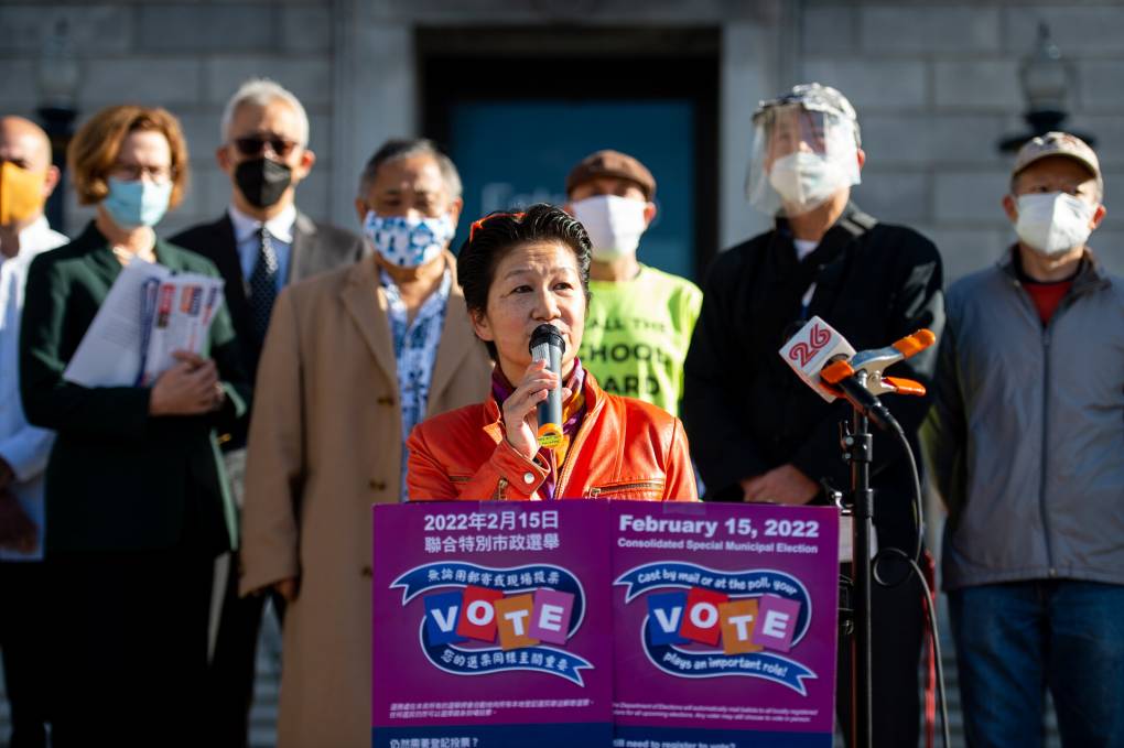 A woman speaks at a podium flanked by other people behind her wearing masks.